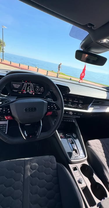 Modern Audi RS3 interior with a digital cockpit and leather steering wheel parked by the ocean.