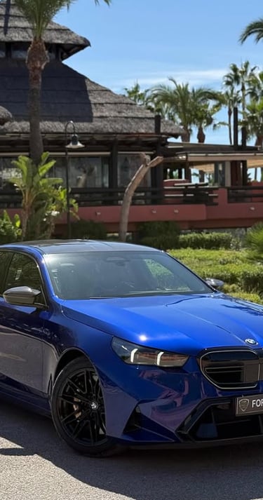 A blue luxury BMW sedan for rent parked in front of a tropical resort with palm trees.