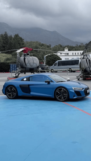 Blue Audi R8 luxury sports car parked on a helipad with black helicopters and mountain scenery.