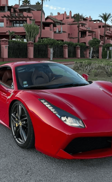 A luxury red Ferrari sports car parked on a road in front of a Mediterranean resort.