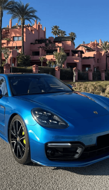 A metallic blue Porsche Panamera parked in front of a luxury pink Mediterranean resort with palm trees.