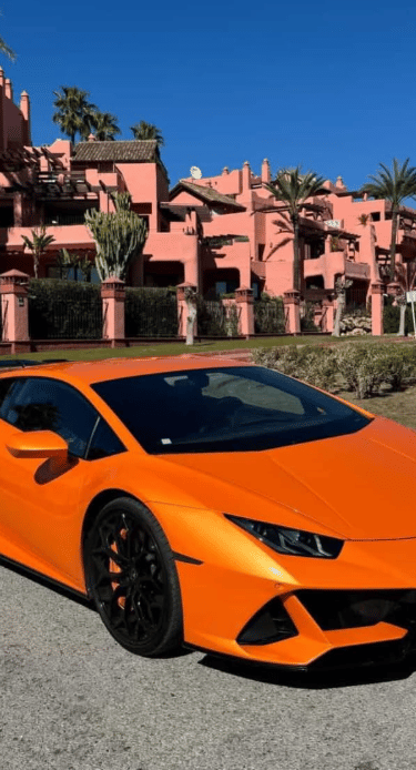 Orange Lamborghini Huracan supercar parked in front of pink Mediterranean villas and palm trees.