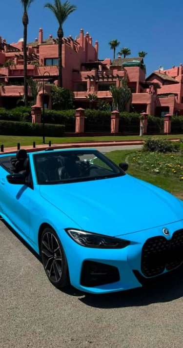 A bright blue convertible BMW parked on a sunny driveway in front of a pink Mediterranean style luxury resort.