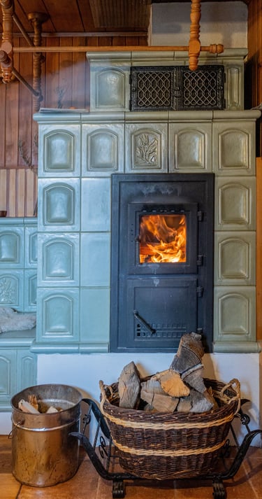Traditional ceramic tile stove with a roaring fire and firewood basket in a cozy wood-paneled room.