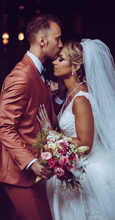 Mariés s'embrassant tendrement : marié en costume terracotta, mariée en robe blanche avec voile.