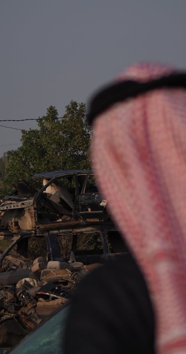 an arab man in a red and white scarf looking at a destroyed car in the by the palm trees in Iraq