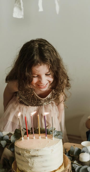 niña sonriente con cabello rizado mira las velas encendidas sobre un pastel de cumpleaños con betún