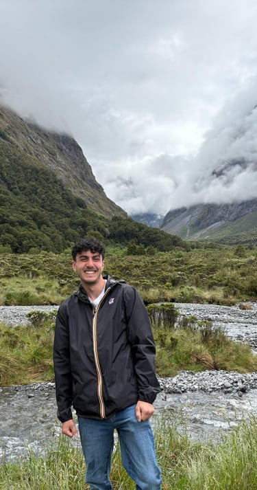 Outdoor portrait of Luca smiling in a mountain valley with a river and cloudy peaks in the backgrou