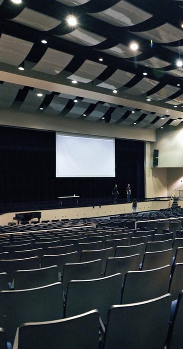 Large empty auditorium with rows of chairs facing a stage and projection screen, under a ceiling wit