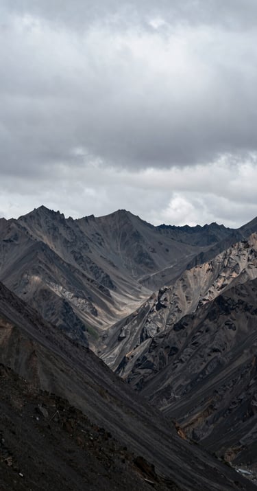 A wide shot of a jagged mountain range under a heavy, overcast soft grey sky. The landscape is desolate and powerful, with high-contrast shadows. Captured in a remote Global / Western mountain pass.