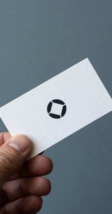 Close up of a professional business card with a minimalist logo, held by a hand in a South American / Brazilian setting, background of a muted steel blue wall.