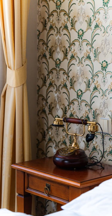 Vintage rotary phone on a wooden nightstand next to floral wallpaper and yellow curtains.