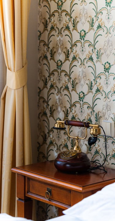 Vintage rotary phone on a wooden nightstand next to floral wallpaper and yellow curtains.