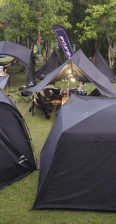 Black dome tents and camping gear set up at a lush outdoor campsite near a forest cabin.