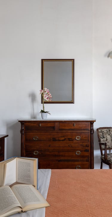Rustic Italian bedroom interior featuring a wooden dresser, floral pillows, and open books on a bed.