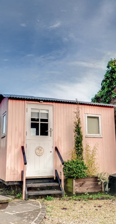 A charming pink shepherd's hut guest room with a wooden picnic table and patio seating.