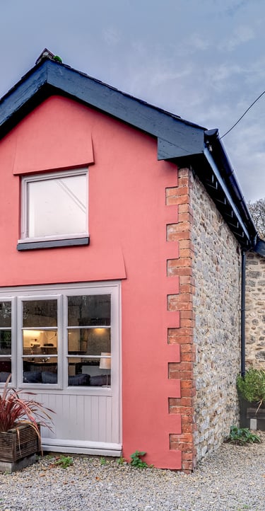 Charming red cottage with stone walls, large windows, and a gravel courtyard under a blue sky.