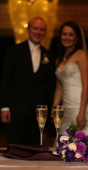 Champagne Glasses and Bridal Bouquet on table in focus. Bride and Groom Looking at camera smiling.