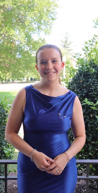 Smiling woman wearing a blue satin cowl neck dress standing outdoors in a garden setting.