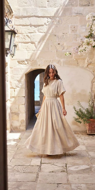 Woman in a beige linen maxi dress walking through a sunlit Mediterranean stone courtyard.