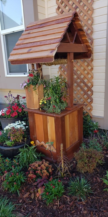 Rustic wooden wishing well planter with a cedar shingle roof in a colorful flower garden.