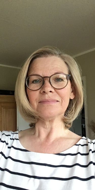 A blonde middle-aged woman wearing round glasses and a striped shirt in a home setting.