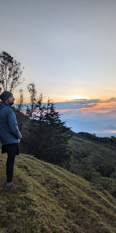 A male hiker standing on a grassy mountain ridge watching a scenic sunrise over a misty valley.