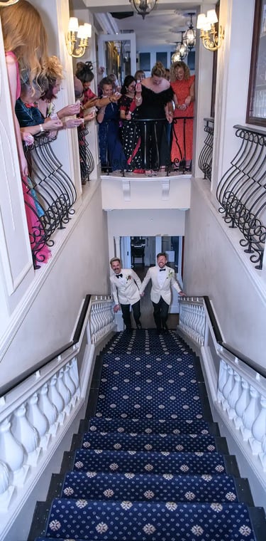 Grooms in white tuxedo jackets walking up a blue carpeted staircase at their wedding reception.
