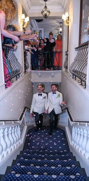 Grooms in white tuxedo jackets walking down stairs as guests throw pink wedding confetti.