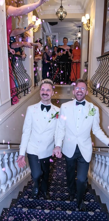 Two grooms in white tuxedo jackets walking down a staircase as guests throw pink flower petal confetti.