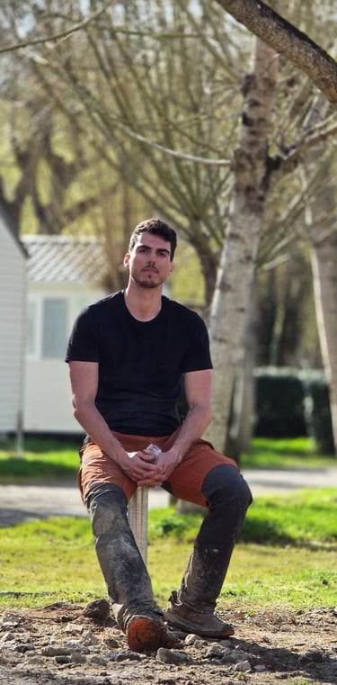 A man in workwear sitting at a sunny campsite with mobile homes and trees in the background.