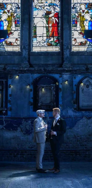 Groom and guest talk inside a rustic wedding venue with stained glass windows and stone walls.