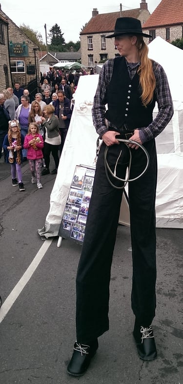 A tall stilt walker performer in a black vest and hat at a crowded outdoor community street festival.