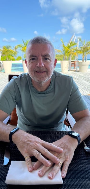 "Person seated at outdoor table in tropical resort with palm trees and lounge chairs nearby