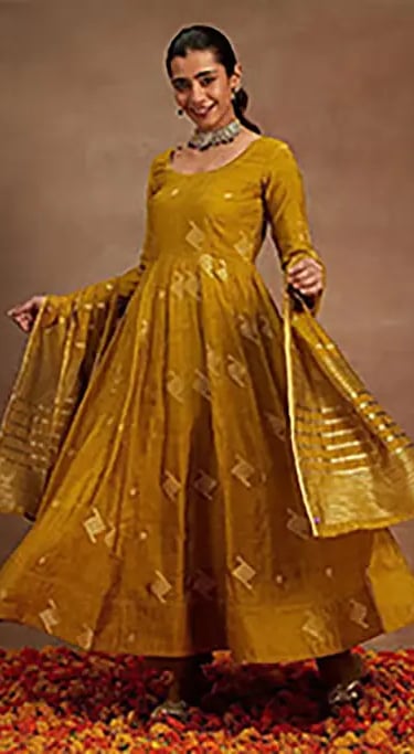A woman posing in a mustard yellow Anarkali suit with a matching dupatta and gold embroidery.