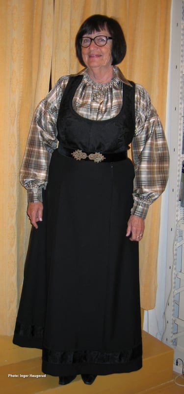 A woman with black hair and glasses stands in a black bunad with a plaid shirt