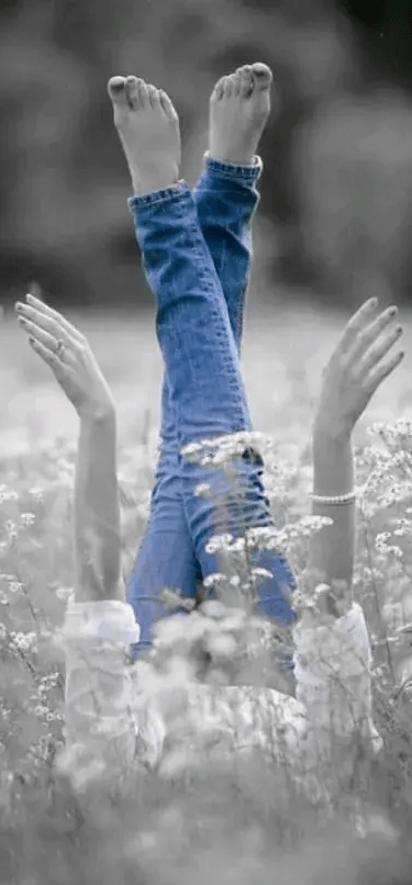 A black and white image of a woman kicking her legs up in a field. The only color is her blue jeans.
