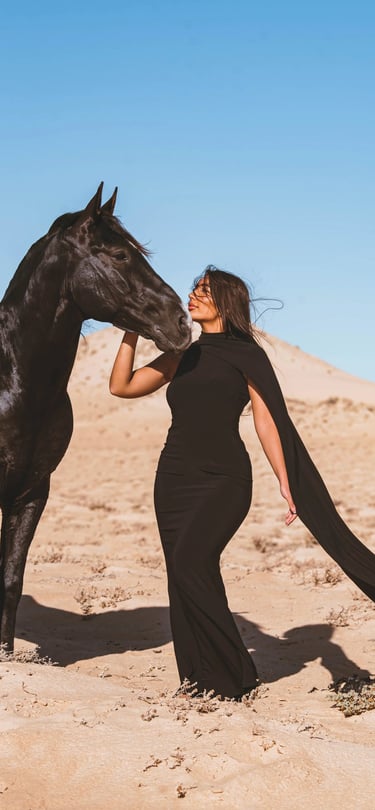 Luxury equestrian fashion shoot in Dakhla featuring a model in a black gown posing with a black horse in the dunes.