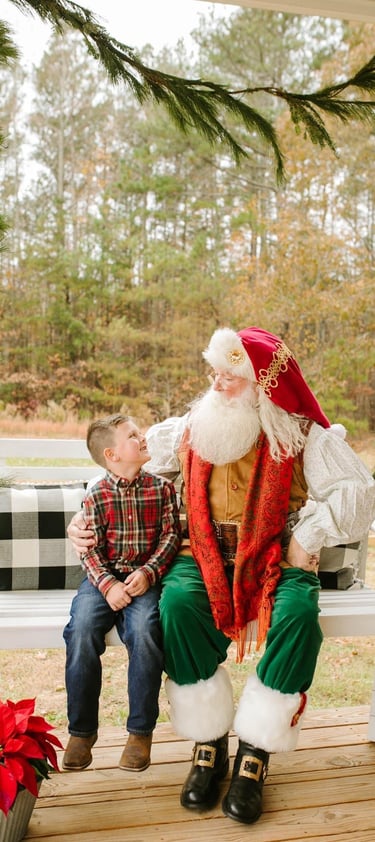 A young boy sits with Santa Claus on a white porch swing at a Christmas tree farm.