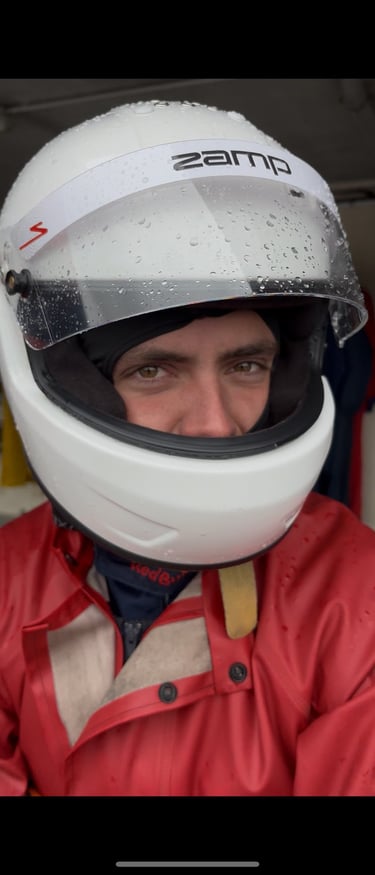 A racing driver wearing a white Zamp helmet and red rain suit with water droplets on the visor.