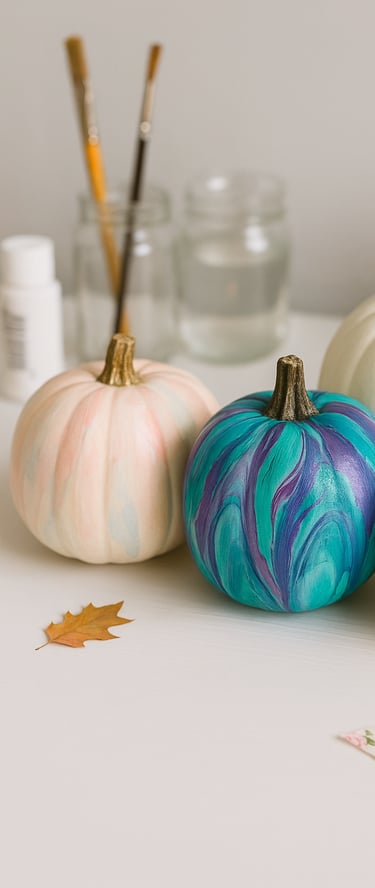Three decorated pumpkins on a white craft table: one with pastel watercolor drips, one with teal 
