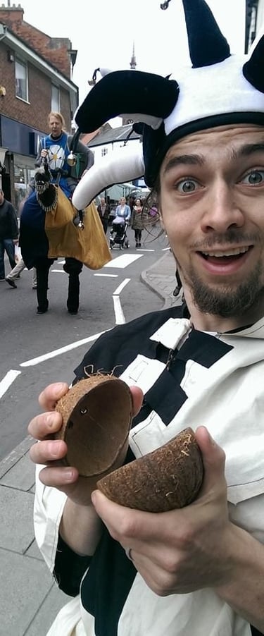 A street performer in a jester hat holding coconut halves for horse sound effects at a festival.