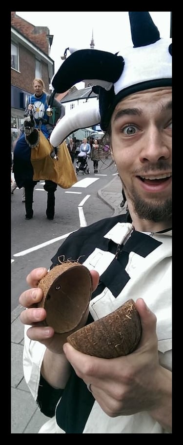 A man in a jester hat holding coconut halves at a Monty Python themed street festival parade.