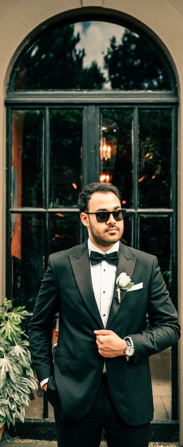 Groom in a black tuxedo and sunglasses standing confidently by a large window, by Fred Art Studio