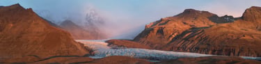 Panoramic sunset view of a massive glacier flowing between rugged volcanic mountains in Iceland.