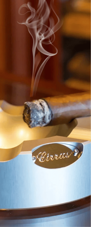 A smoking cigar rests in a wooden ashtray, with smoke curling upwards, suggesting a strong odor.