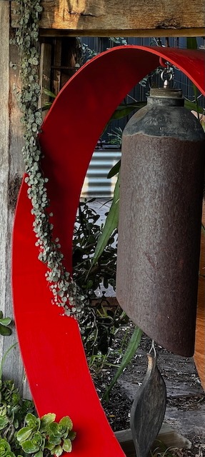 Rustic metal wind chime bell hanging inside a red circular garden sculpture with green vines.