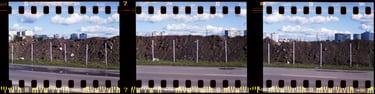 Composed photo featuring a street with a prominent, dark, rocky mound, with the cityscape in the background and clouds on sky