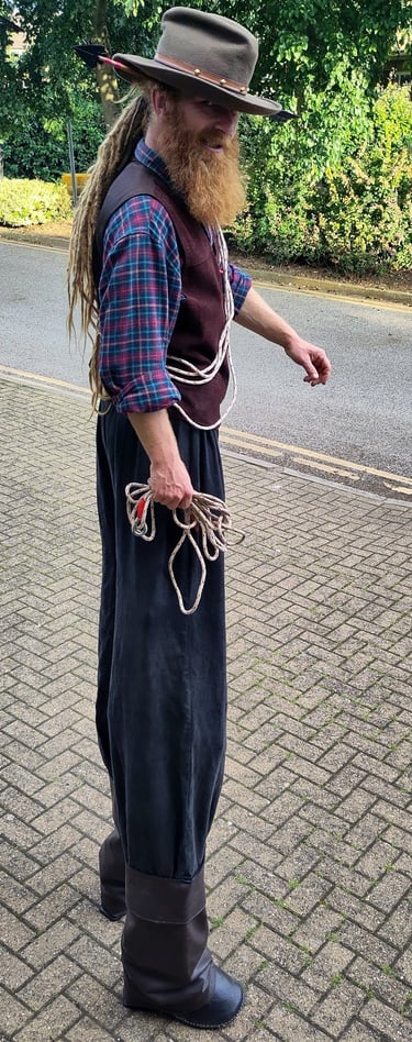 A stiltwalker with a long ginger beard wearing a cowboy hat and western attire holding a lasso