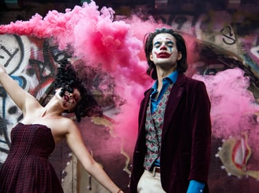 A model dressed as the Joker poses while a Harley Quinn spreads purple smoke in the background